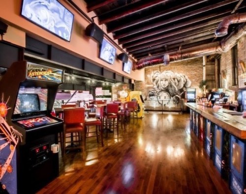 A retro arcade with vintage games, bar seating, and a mural on the brick wall, featuring multiple screens overhead.