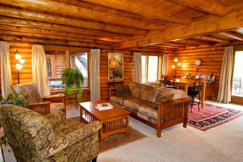 Cozy log cabin interior with wooden beams, comfortable seating, a dining area, and warm lighting.