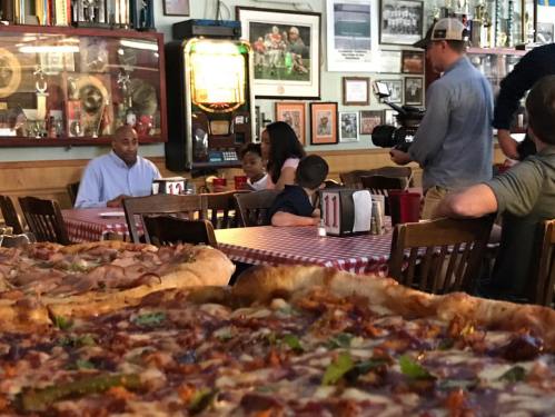 A busy pizzeria with large pizzas in the foreground and diners, including a family and a person filming, in the background.