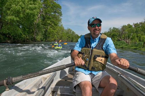 A man in a boat navigates a river, wearing sunglasses and a life vest, with kayakers visible in the background.