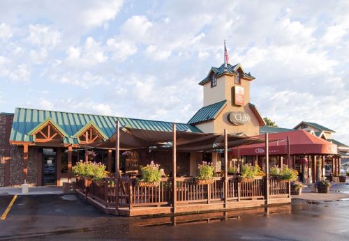 A rustic building with a green roof, featuring outdoor seating and flower planters, under a blue sky with clouds.