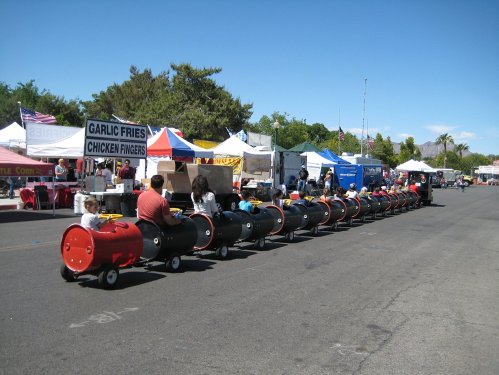 A line of children riding a small train made of barrels at a fair, with food stalls and colorful tents in the background.