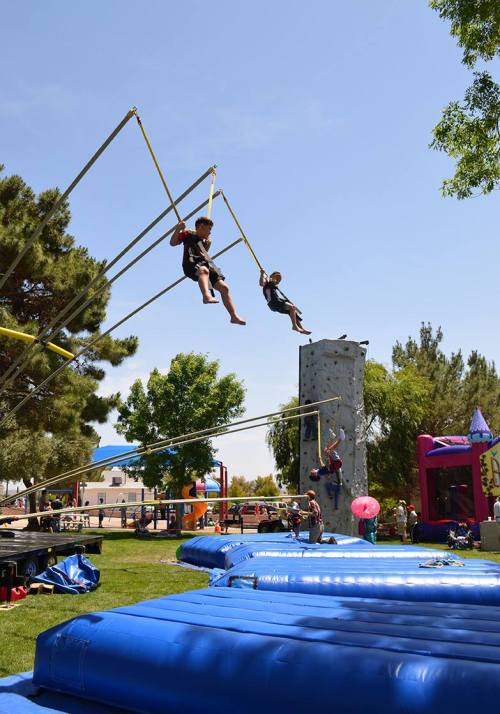 Two children swing on a bungee trampoline while another climbs a rock wall in a sunny park setting.