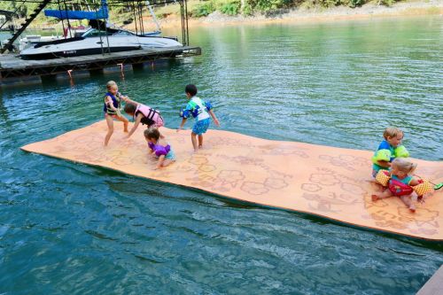 Children playing on a floating mat in a lake, wearing life jackets, with boats in the background.
