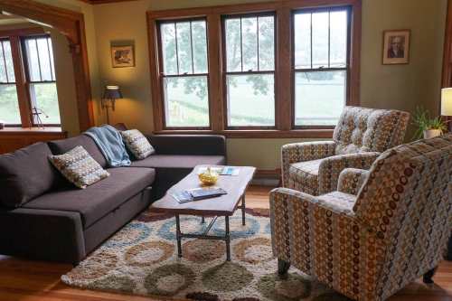 Cozy living room with a sofa, patterned chairs, a coffee table, and large windows overlooking a green landscape.