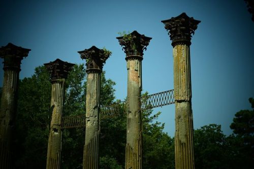Ancient stone columns with ornate capitals stand amidst greenery, with a bridge connecting some of them.