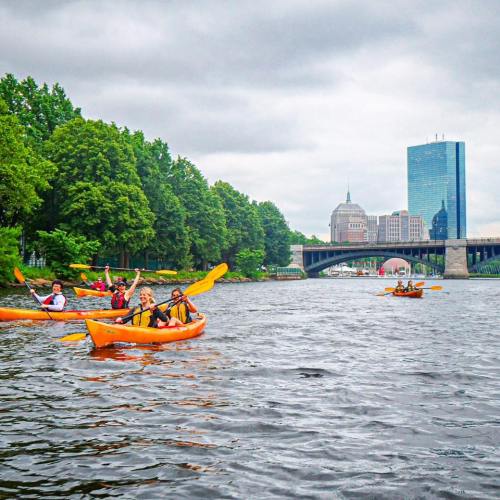 A group of people kayaking in orange kayaks on a river, with a city skyline and green trees in the background.