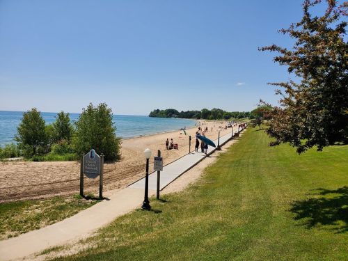 A sunny beach scene with people enjoying the sand and water, surrounded by greenery and a walking path.
