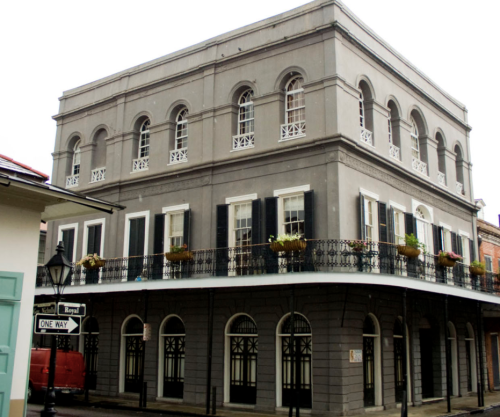 Historic gray building with arched windows and a balcony, adorned with flower pots, located on a street corner.