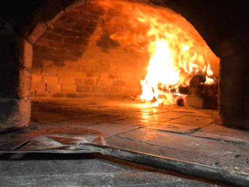 A view inside a brick oven with flames and glowing embers, featuring a metal pizza peel in the foreground.