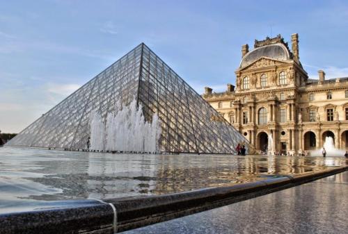 The Louvre Museum's glass pyramid reflects in the water, with the historic building in the background under a clear sky.