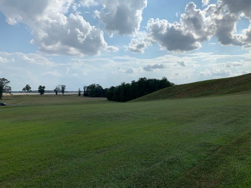 A wide grassy field under a partly cloudy sky, with trees in the distance and a gentle slope on one side.