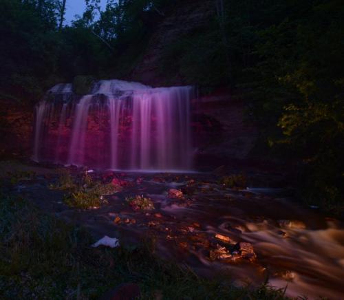 A serene waterfall illuminated in purple light, surrounded by lush greenery and flowing water at dusk.