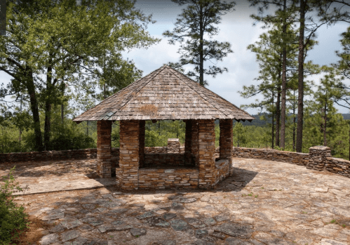 A stone gazebo with a wooden roof, surrounded by trees and a scenic view of the landscape.