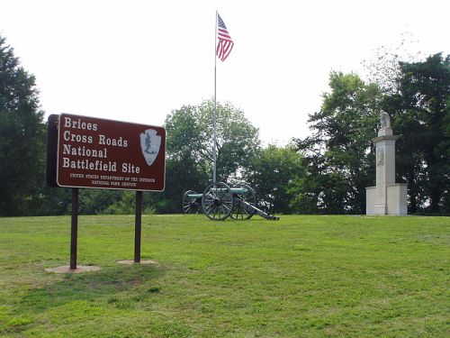 Sign for Brices Cross Roads National Battlefield Site with a cannon and monument in the background, under an American flag.