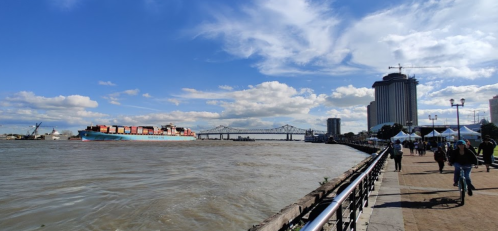 A riverfront scene with a cargo ship, people walking, and buildings under a blue sky with scattered clouds.