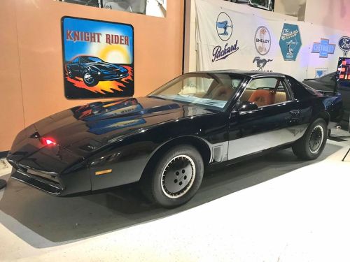 A black sports car with a red light on the front, displayed in a museum with automotive memorabilia in the background.