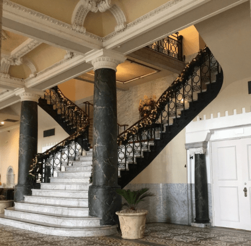 Elegant interior featuring a grand staircase with ornate railings, marble columns, and decorative holiday garlands.