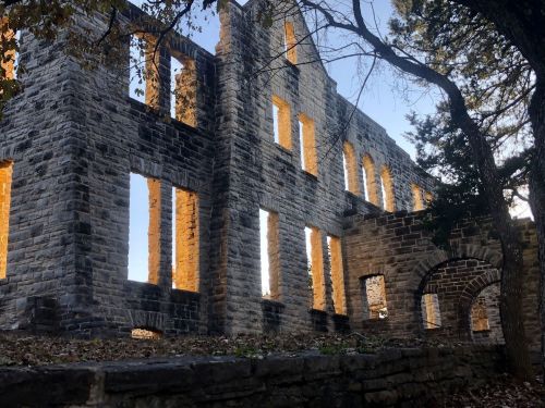 Ruins of a stone building with arched windows, surrounded by trees and a clear blue sky.