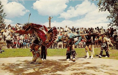 Indigenous dancers in traditional attire perform in front of an audience outdoors, with trees and clouds in the background.