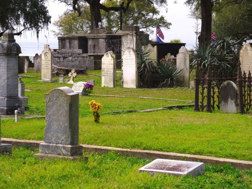 A serene cemetery with various gravestones, surrounded by grass and trees, featuring American flags in the background.