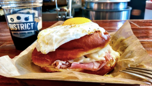 A sandwich with ham, melted cheese, and a fried egg on a bun, alongside a cup of iced coffee.