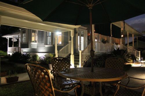 A well-lit house at night with a patio table and chairs under an umbrella, surrounded by greenery.