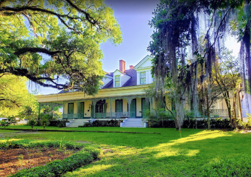 A large, historic southern mansion surrounded by lush greenery and trees, featuring a wraparound porch and Spanish moss.