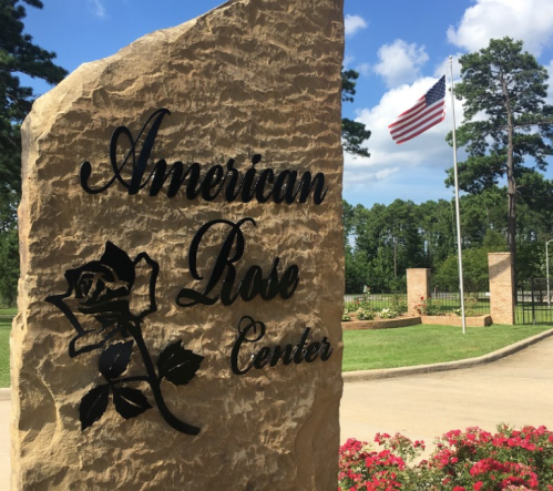 Sign for the American Rose Center with a rose design, surrounded by greenery and an American flag in the background.
