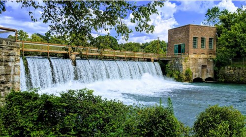 A scenic view of a waterfall cascading over a dam, with a brick building nearby and lush greenery surrounding the area.