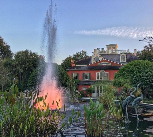 A vibrant fountain sprays water in front of a red mansion, surrounded by lush greenery and decorative sculptures.