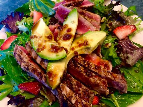 A colorful salad topped with grilled steak, avocado slices, strawberries, and a drizzle of dressing.