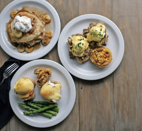 Three plates of food: pancakes with syrup, eggs Benedict with corn, and asparagus on a wooden table.