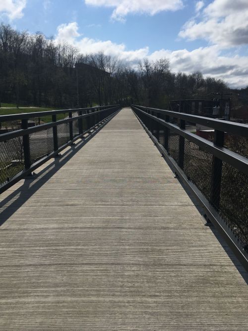 A wide, empty pedestrian bridge stretches into the distance, surrounded by trees and a cloudy sky.
