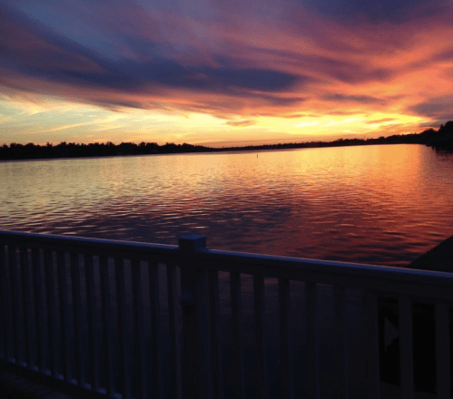 A vibrant sunset over a calm lake, with colorful clouds reflecting on the water and a white railing in the foreground.