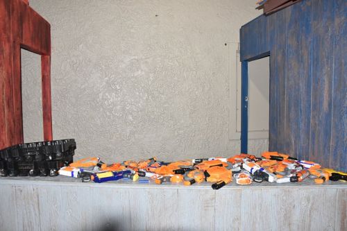 A table covered with various toy blasters in orange and blue, set against a textured wall and colorful wooden structures.