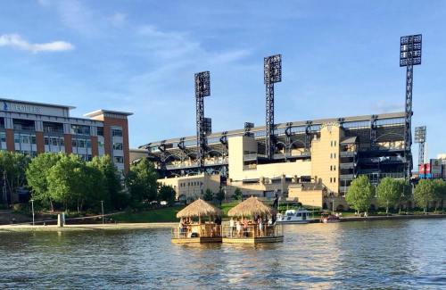 A scenic view of a stadium by the water, featuring a tiki hut on the lake and lush greenery along the shore.
