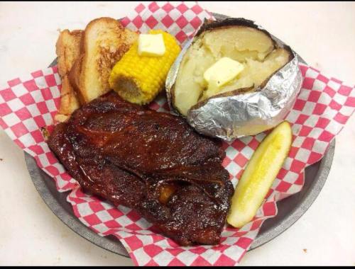 A plate with barbecue meat, a baked potato with butter, corn on the cob, toast, and a pickle slice on a checkered paper.