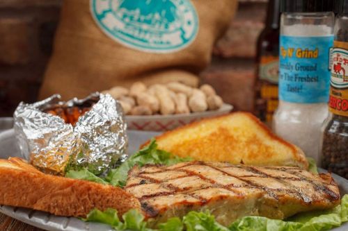 Grilled fish fillet on lettuce with toast, foil-wrapped potatoes, peanuts, and condiments in the background.