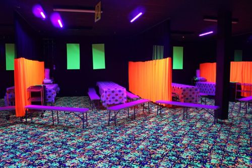 A brightly lit room with neon curtains, patterned tables, and a colorful floor, creating a vibrant, playful atmosphere.