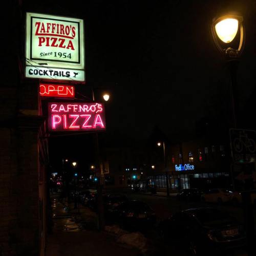 Neon signs for Zaffiro's Pizza and cocktails illuminate a street at night, with nearby buildings and streetlights visible.