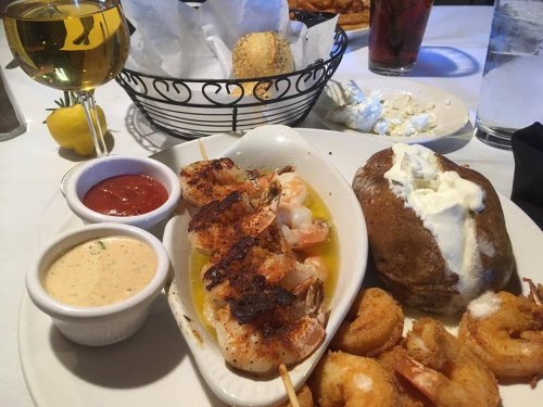 A plate featuring grilled shrimp, a baked potato with sour cream, and fried shrimp, accompanied by sauces and drinks.