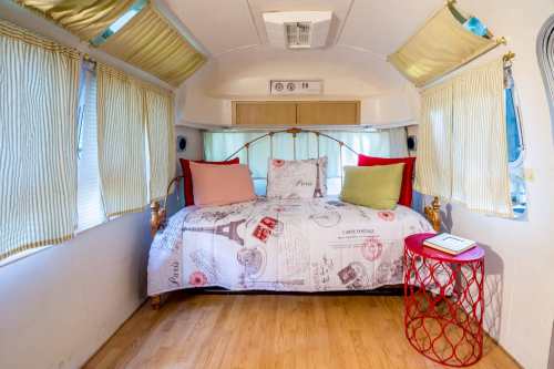 Cozy interior of a vintage Airstream trailer with a bed, decorative pillows, and a small red table. Natural light from windows.