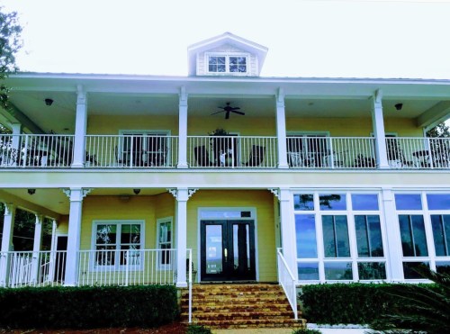 A large, two-story yellow house with a balcony, white railings, and large windows, surrounded by greenery.