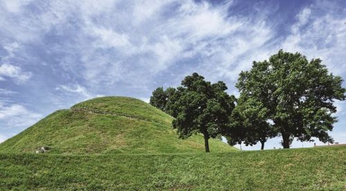 A grassy hill with a stone path, surrounded by trees under a blue sky with scattered clouds.
