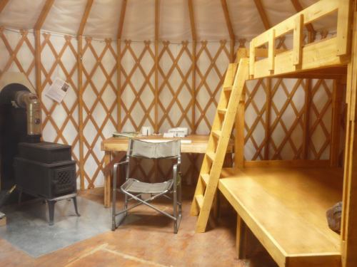 Interior of a yurt featuring a wooden bunk bed, a chair, a table, and a small stove. Warm, rustic decor.