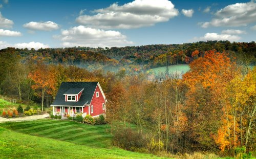 A red house surrounded by vibrant autumn trees and rolling hills under a blue sky with fluffy clouds.
