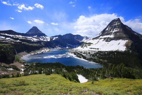 A stunning mountain landscape featuring snow-capped peaks, a blue lake, and lush green meadows under a bright sky.