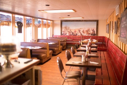 Cozy restaurant interior with wooden booths, tables, and a mural of cowboys on horseback. Warm, inviting atmosphere.