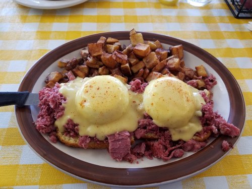 A plate of eggs Benedict with corned beef hash, topped with hollandaise sauce, served with crispy potato cubes.